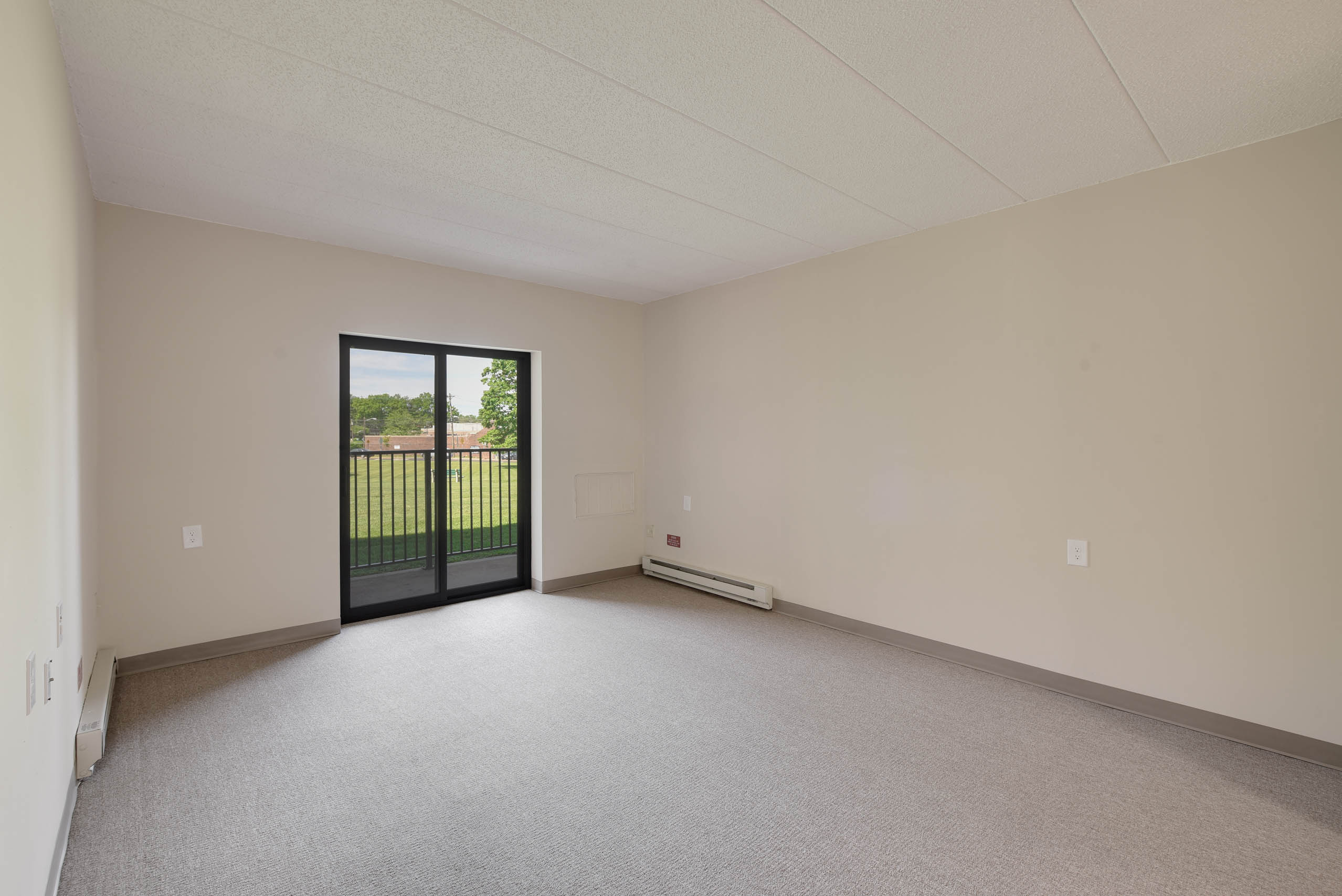 A room with a carpeted floor, a window with a view of a fence, and a door leading outside.