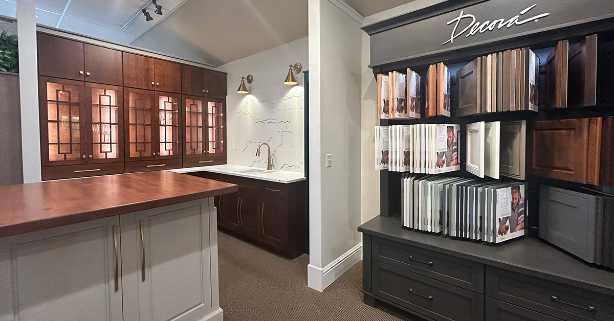 Showroom kitchen vignette with Decora cabinetry, including a center island with a wood countertop, dark wood cabinets with decorative glass panels, wall-mounted lighting, and a nearby display of cabinet door samples.