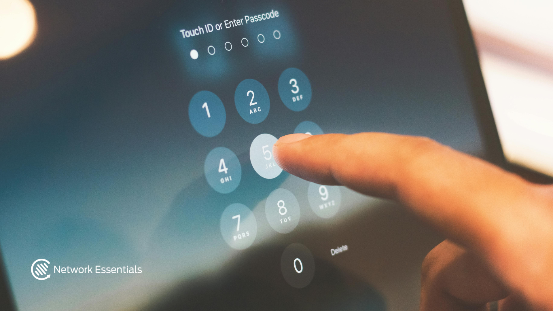 A close-up shot shows a finger pressing the '5' key on a digital keypad, likely on a tablet or smartphone. The screen displays a passcode entry interface with numbers arranged in a grid and the text 'Touch ID or Enter Passcode' at the top. A small logo with the text 'Network Essentials' is visible in the bottom left corner.