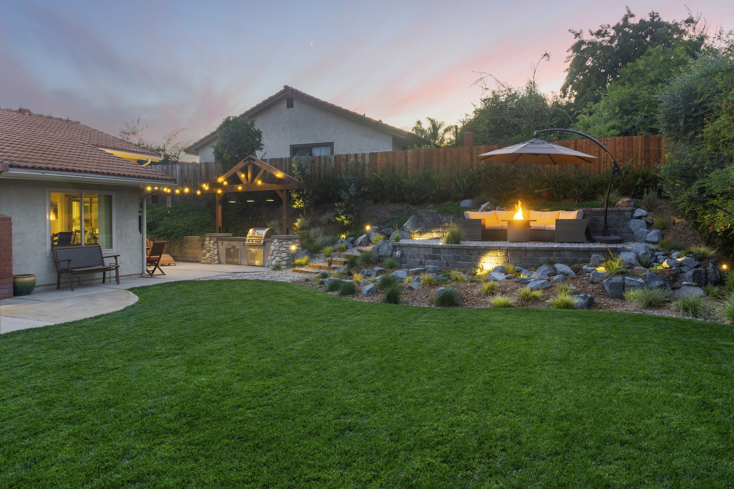 Serene backyard at dusk with lush lawn, stone waterfall, glowing fire feature, patio seating, string lights, and a pastel sunset sky.