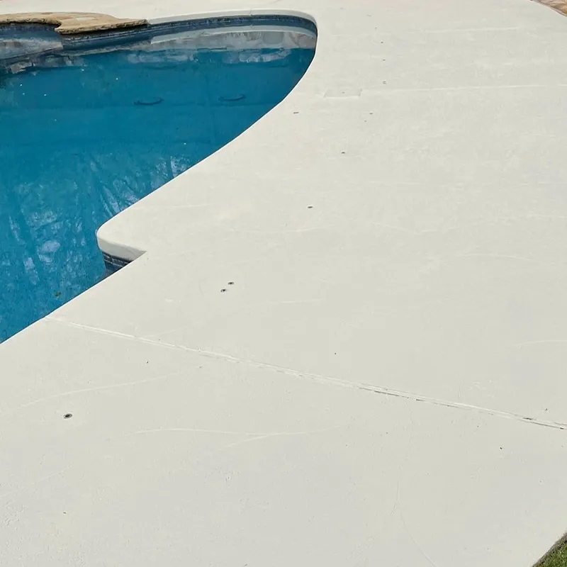 A high-angle view of a light-colored concrete pool deck adjacent to a bright blue swimming pool. The pool's edge is lined with mosaic tiles. The concrete surface has visible seams and some small dark specks.