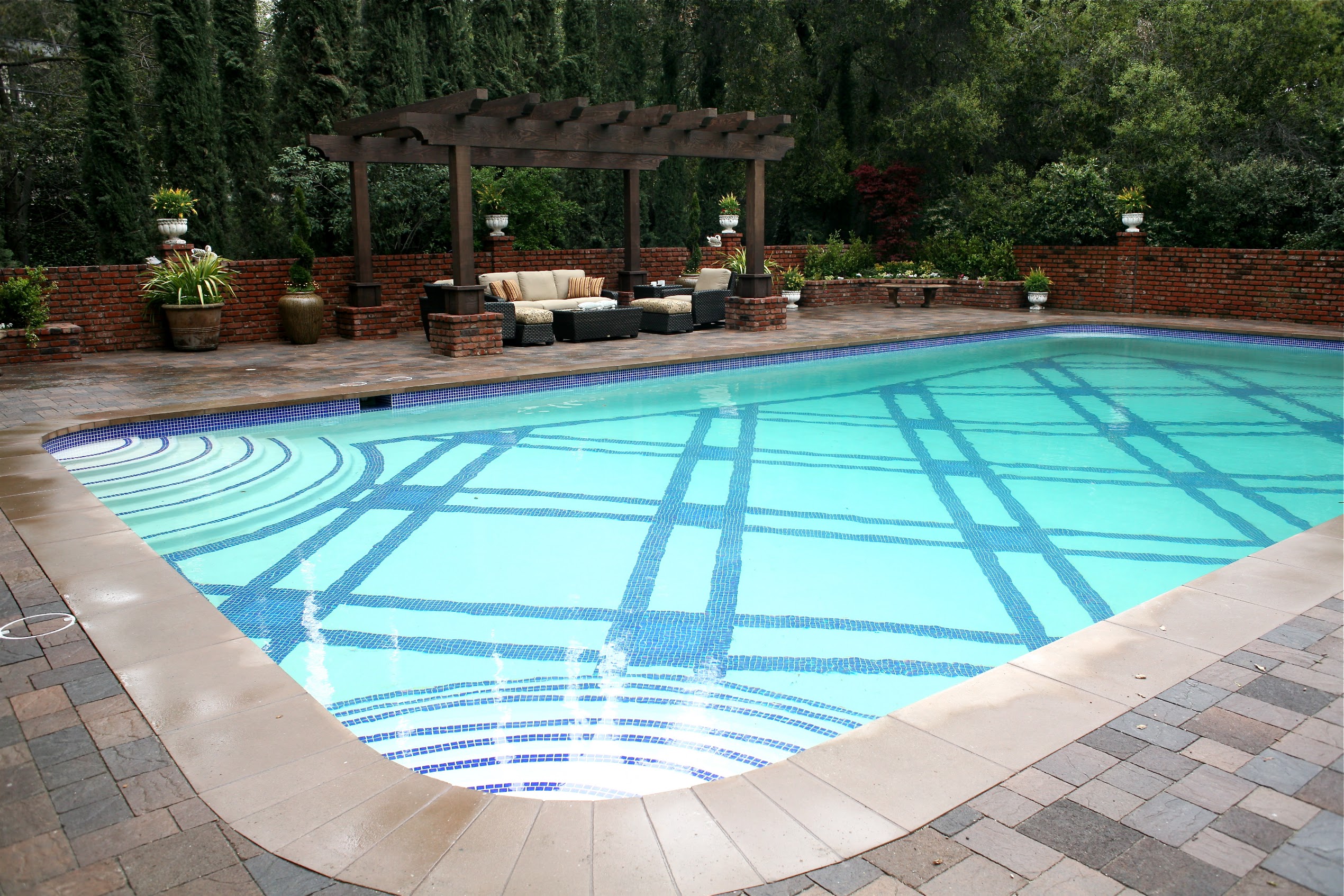 Serene backyard swimming pool with geometric blue tile patterns, stone patio, brick walls, lush greenery, and shaded pergola seating.