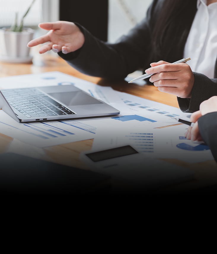 Close-up of two people working together at a desk with an open laptop and documents spread across it.