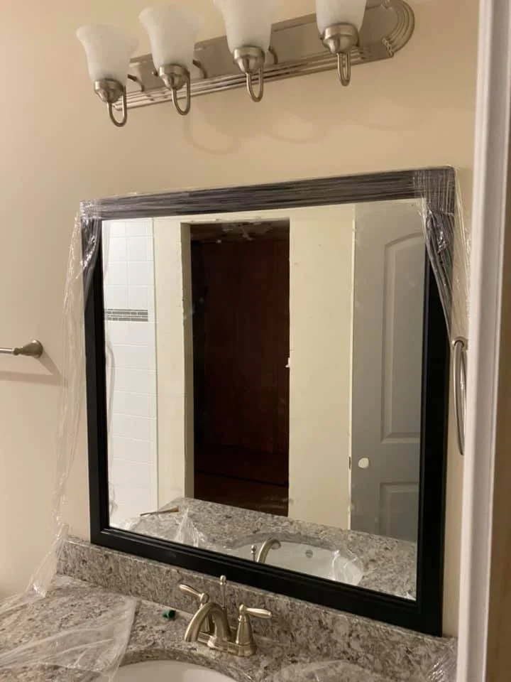 A bathroom vanity with a large framed mirror above it, a sink with a faucet, and a vanity light fixture with four bulbs. The mirror is covered in plastic protective wrap. The countertop appears to be granite or a similar material. The walls are painted a neutral color. The overall scene suggests a recently renovated or new bathroom.