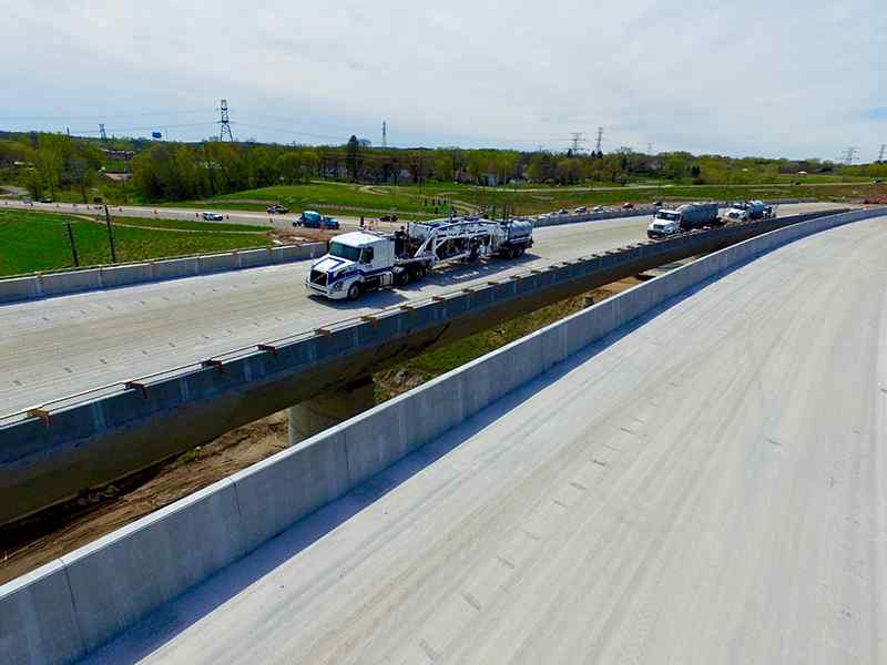Mid-distance aerial perspective of a DSI grinding rig staged on a multi-span concrete highway bridge deck. Background features rolling green fields, power lines, and distant traffic, highlighting active deck preparation work on a major overpass.