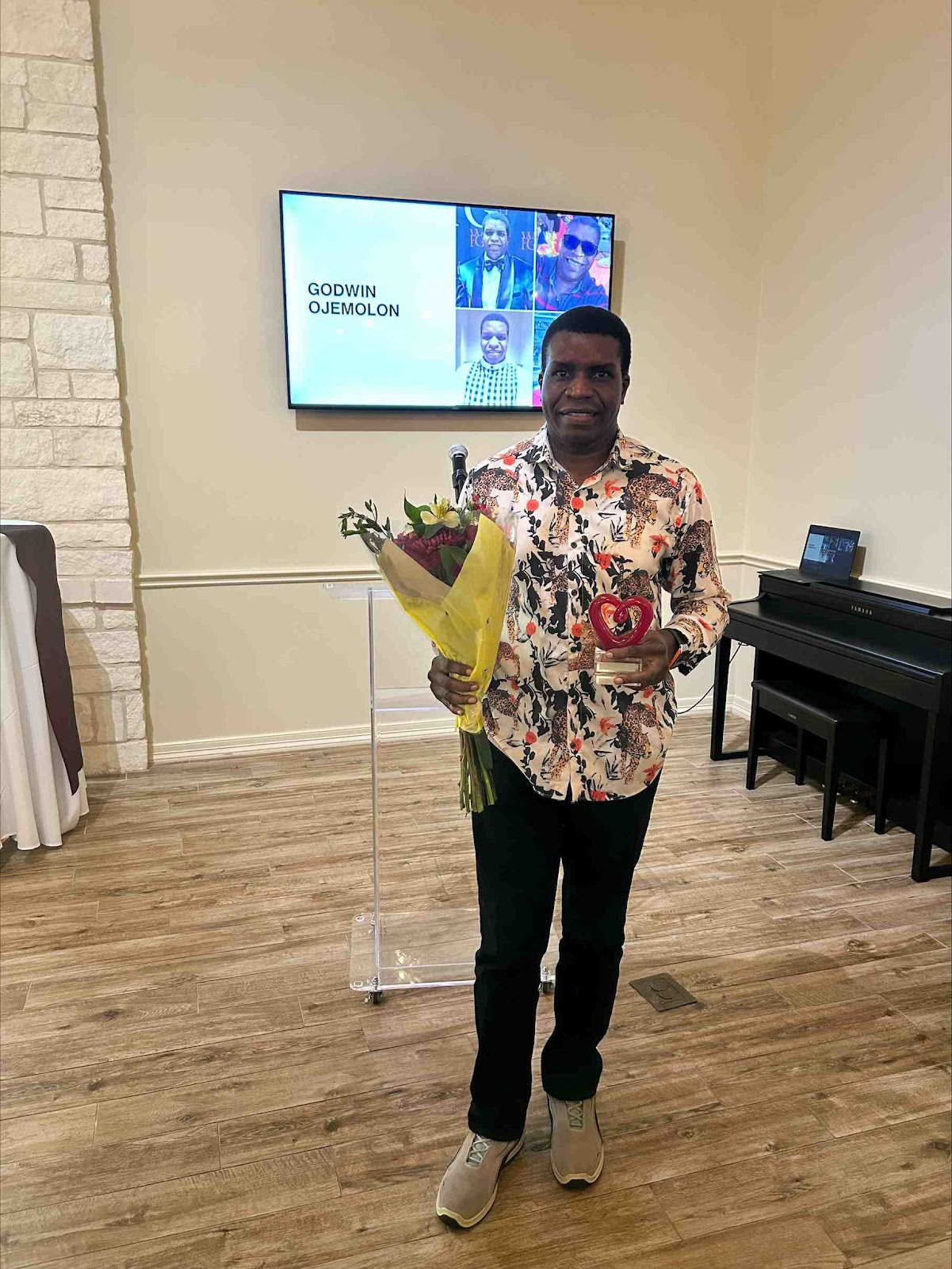 A man stands indoors holding a bouquet of flowers and a heart-shaped award. He is wearing a patterned shirt and dark pants. Behind him, a large screen displays his name, "GODWIN OJEMOLON," along with several smaller images of him. A digital piano with a bench is to his right.