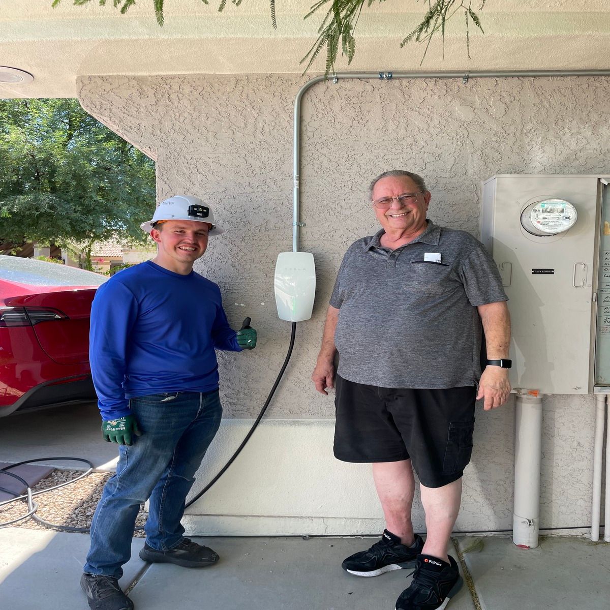 An electrician and homeowner stand smiling beside a newly installed Tesla wall charger mounted on an exterior stucco wall, with electrical conduit, utility equipment, and a red vehicle nearby outdoors.