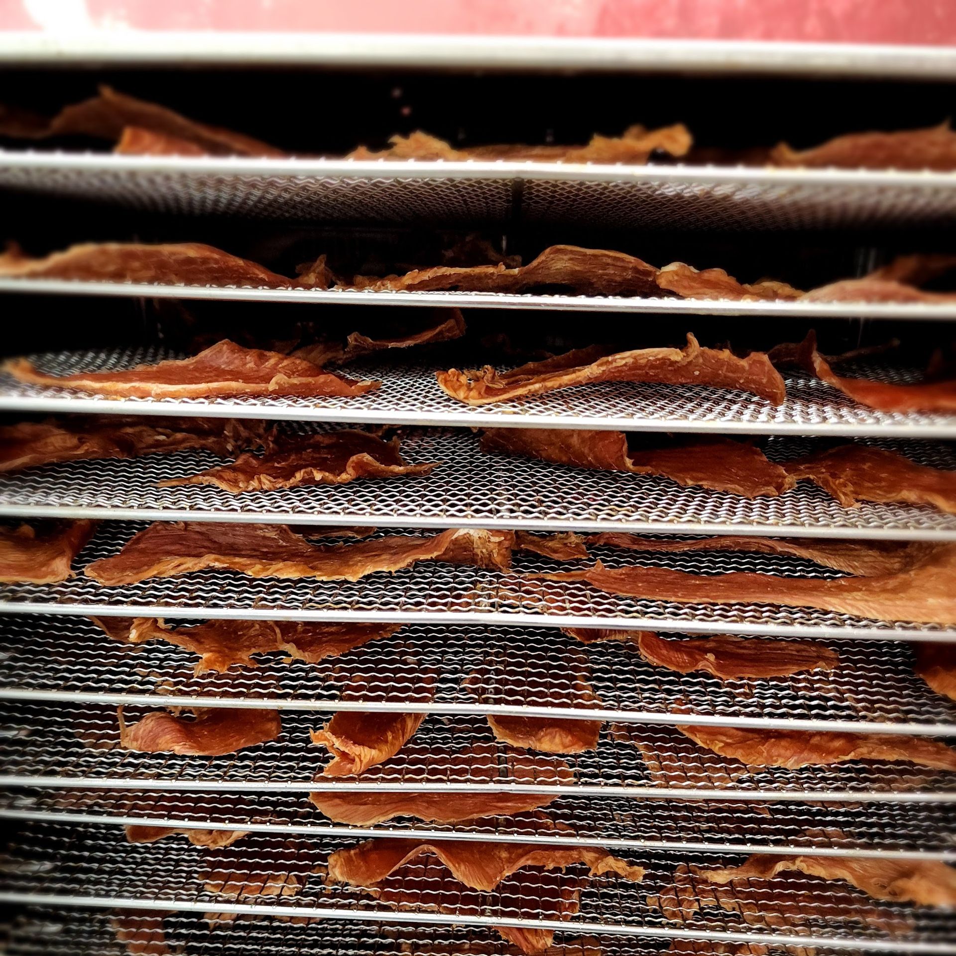Thin slices of meat are arranged on multiple metal trays inside a food dehydrator. The trays are stacked evenly, allowing warm air circulation while the meat slowly dries.