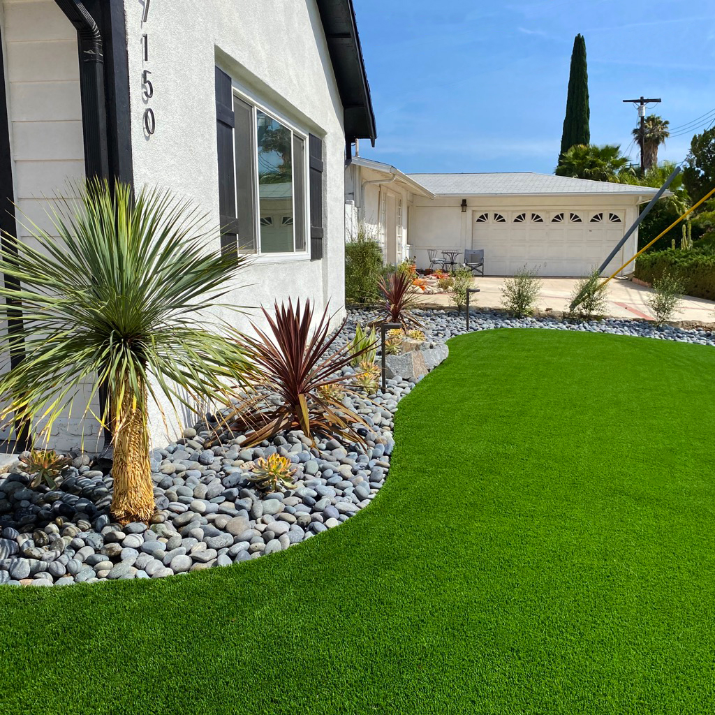 Modern desert-style home with bright artificial turf, river-rock landscaping, yucca and red plants, driveway, and sunny blue skies.