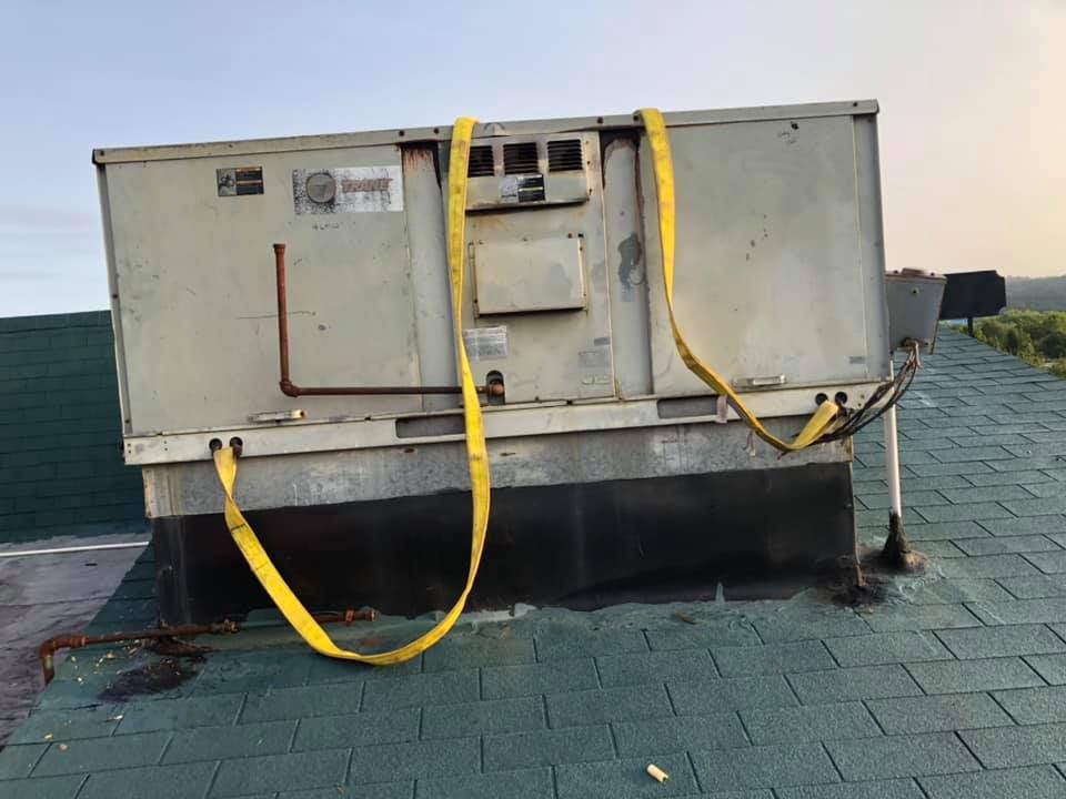 Old rooftop HVAC unit secured with yellow straps, showing rust, wear, and aging components, installed on sloped roof surface, indicating need for repair, maintenance, or replacement in commercial building system.