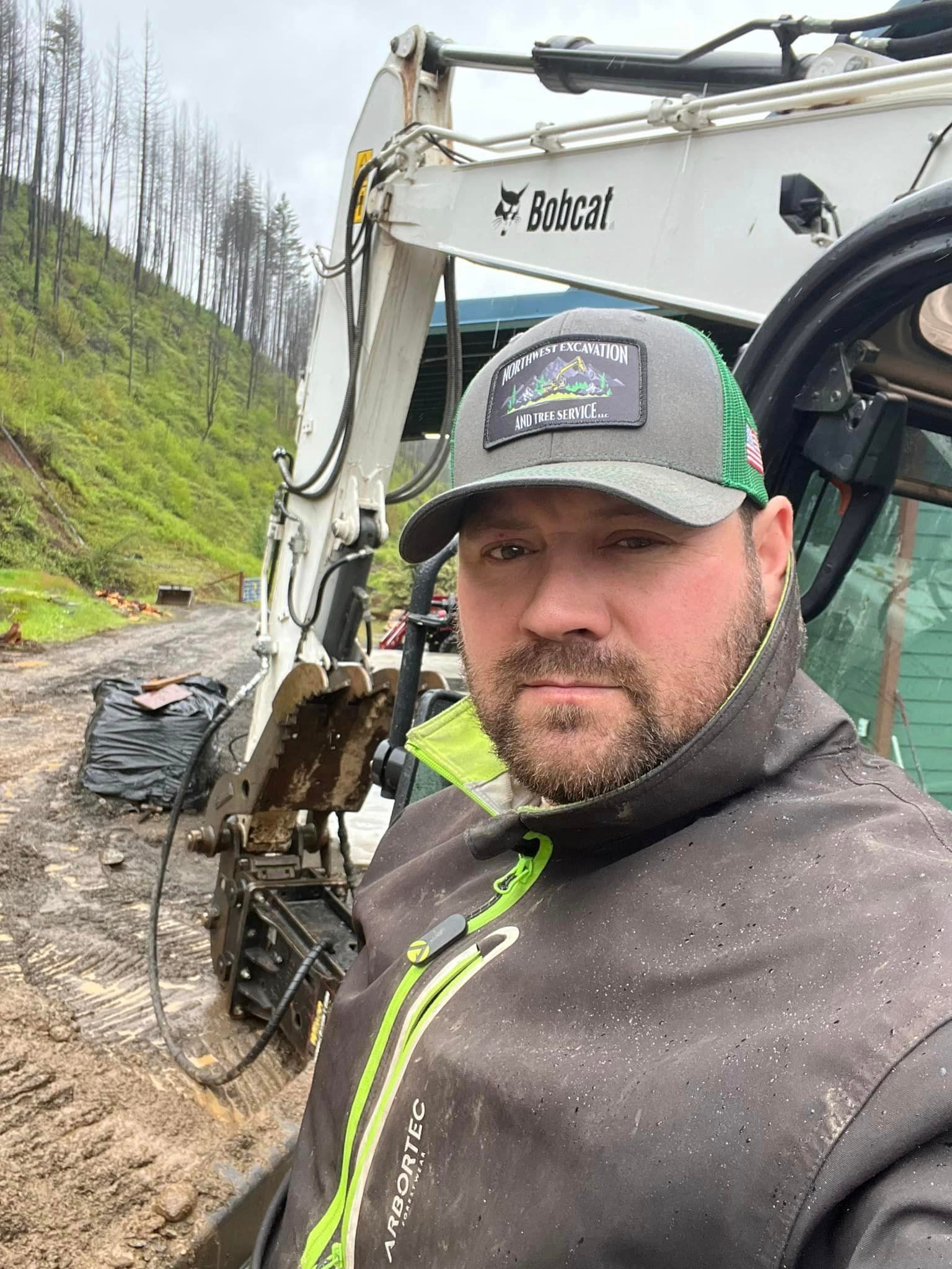 A bearded man in a Northwest Excavation and Tree Service cap and an Arbortec jacket takes a selfie beside a white Bobcat excavator with a hydraulic attachment on a muddy road.