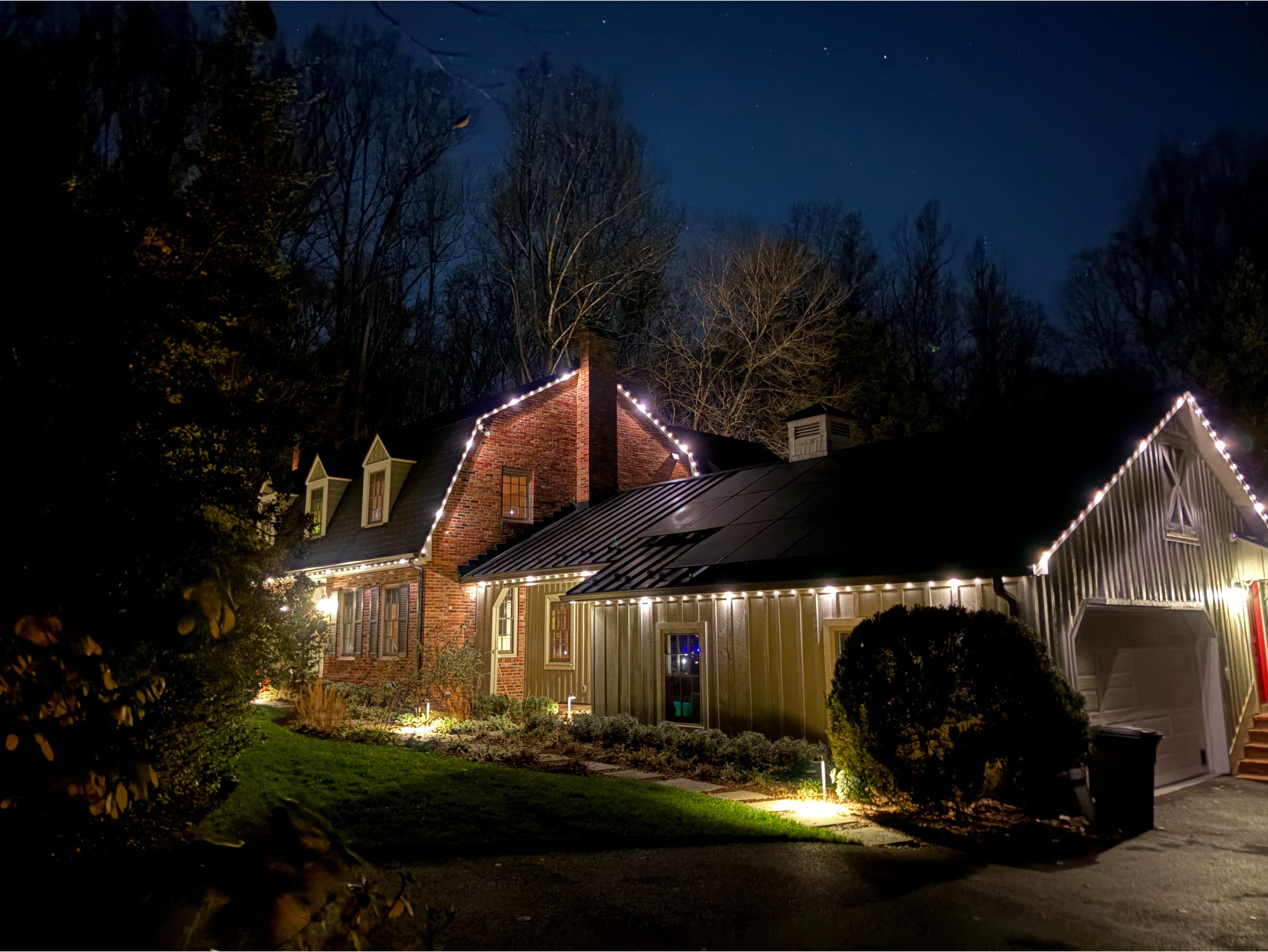 Home at dusk with warm white roofline lighting and landscape lights set among surrounding trees. 