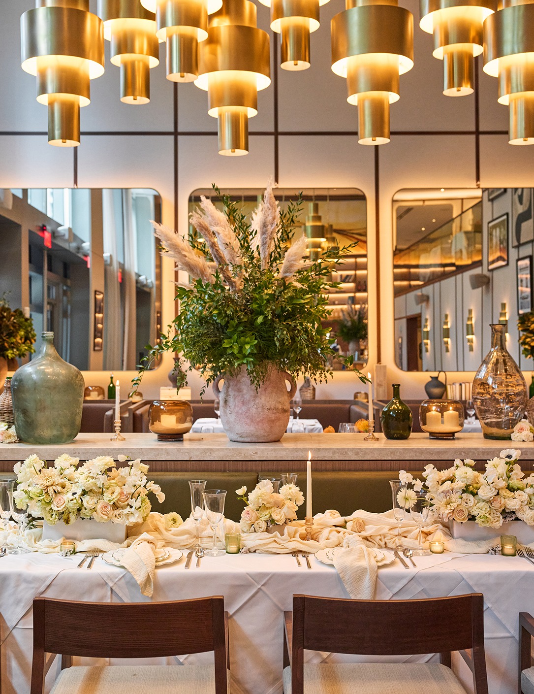 A beautifully set table for a group dinner under tiered gold pendant lights, featuring white roses and pampas grass centerpieces.