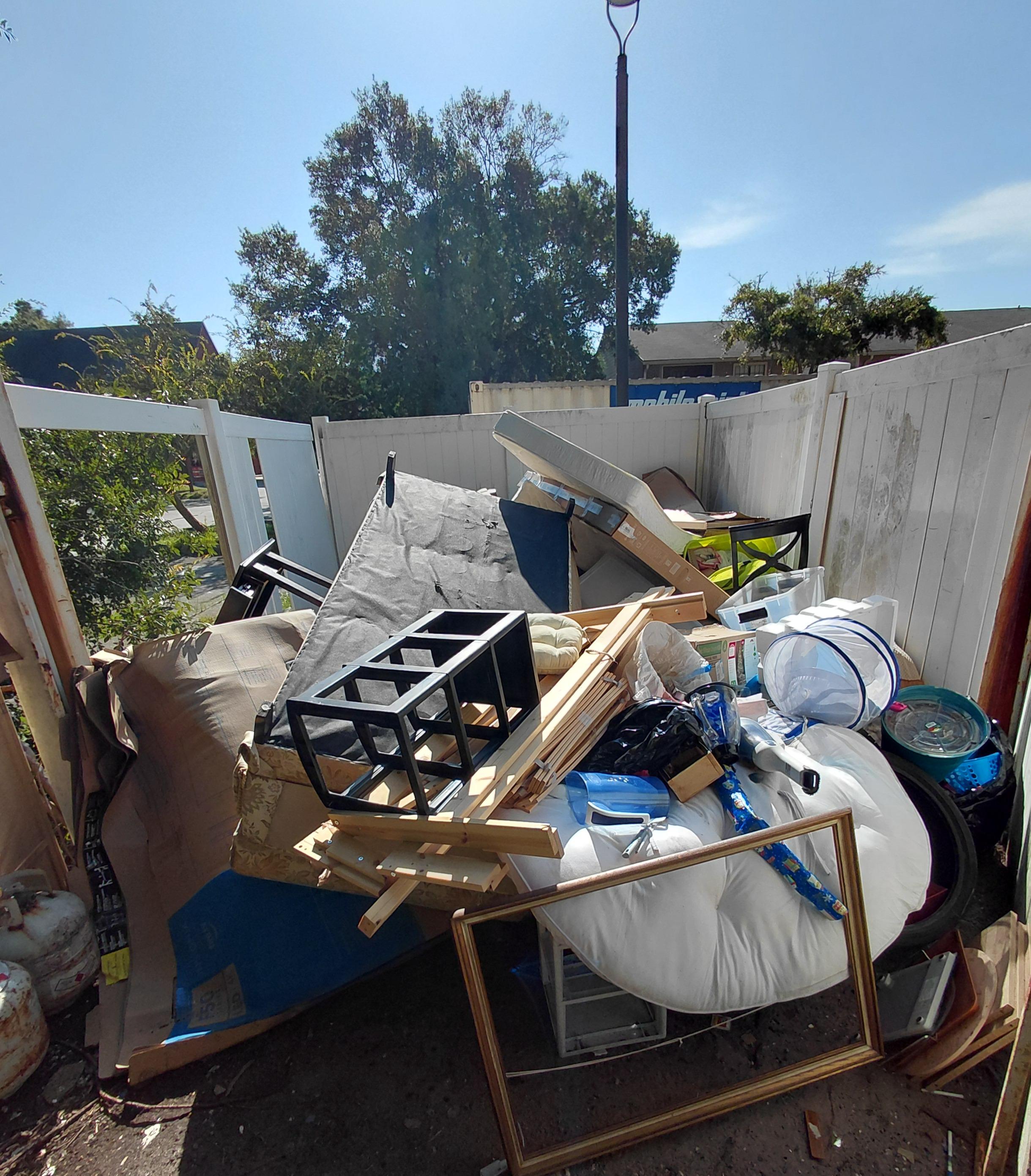A gated outdoor enclosure overflowing with discarded household items, including a box spring, pictur 