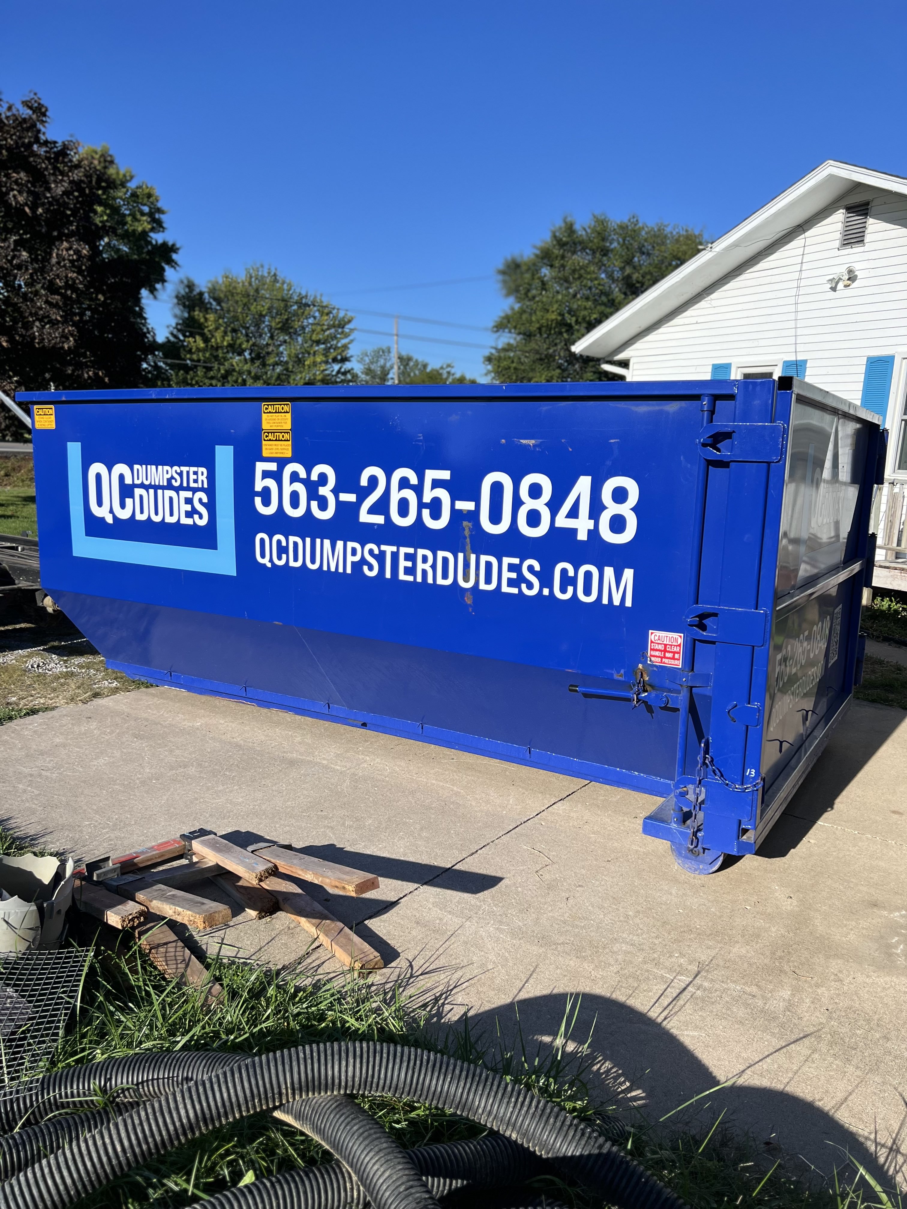 A blue QC Dumpster Dudes dumpster placed in a residential driveway, demonstrating its utility for home cleanouts and light construction waste.