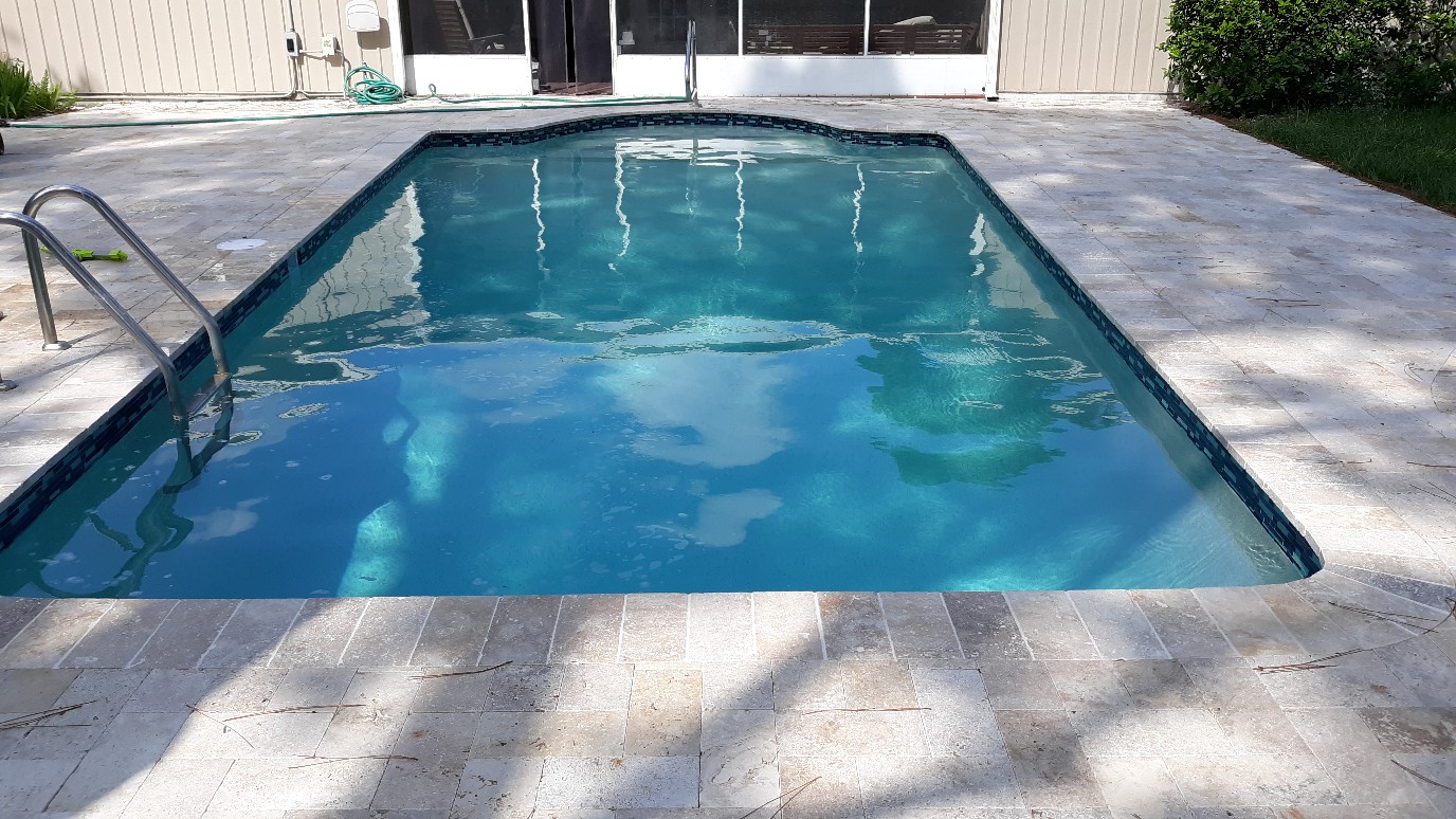 Rectangular residential swimming pool with clear blue water, metal handrails, and light stone decking beside the home.