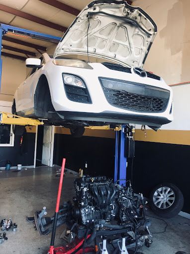 White car lifted on hydraulic hoist inside garage with engine removed below. The scene highlights major mechanical repair or replacement work, demonstrating professional automotive service environment with tools, equipment, and technician workspace clearly visible.
