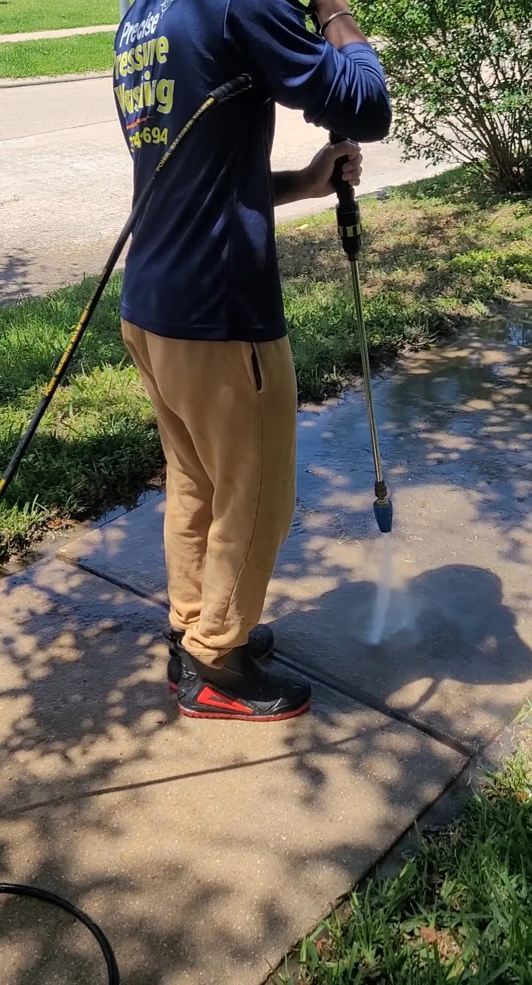 A person in a blue long-sleeved shirt and tan sweatpants is pressure washing a concrete walkway on a sunny day. The person is wearing black and red boots and holding a pressure washer wand, directing a stream of water onto the ground. The walkway is wet and shows signs of being cleaned. Green grass and a leafy bush are visible on the sides of the walkway.