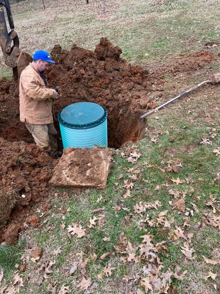 A service technician in a tan jacket and blue hat works on a teal septic tank riser installed in a deep excavation. A yellow excavator and a shovel are nearby.