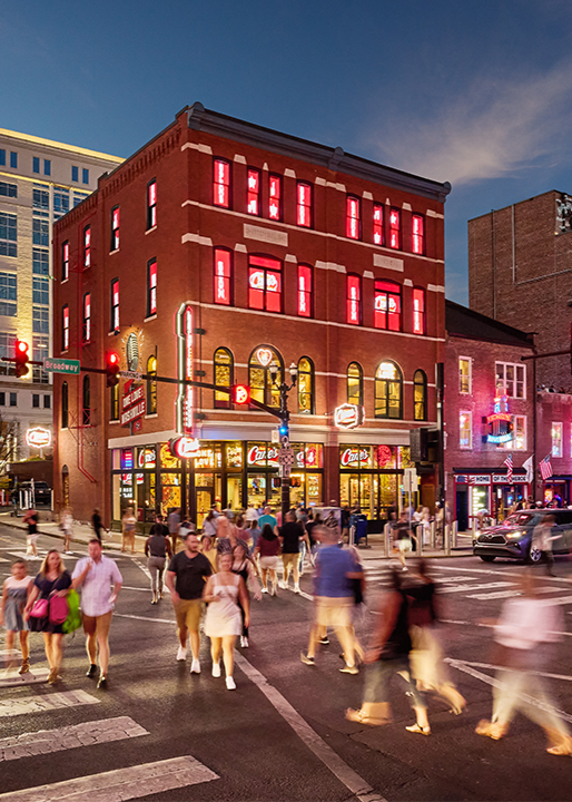 The exterior of a multi-story brick building on Broadway at twilight, illuminated by red lights in the windows and Raising Cane's signage, with a long-exposure effect showing a busy crowd of pedestrians crossing the street in the foreground.