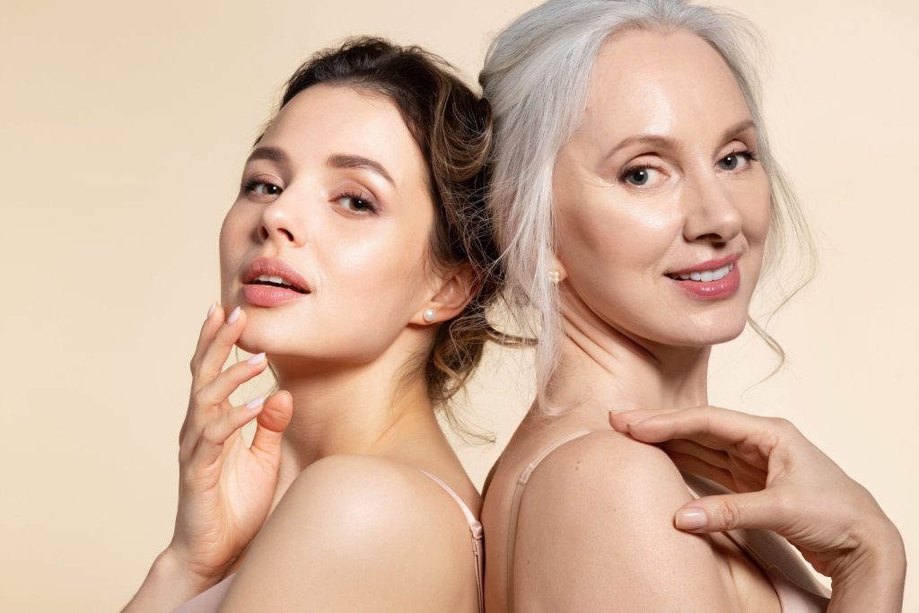 A younger woman with dark hair and an older woman with grey hair stand back-to-back in a studio portrait. Both women are wearing subtle makeup and pearl earrings. The younger woman is touching her chin and neck, while the older woman has her hand on her shoulder. The background is a solid, light beige color.