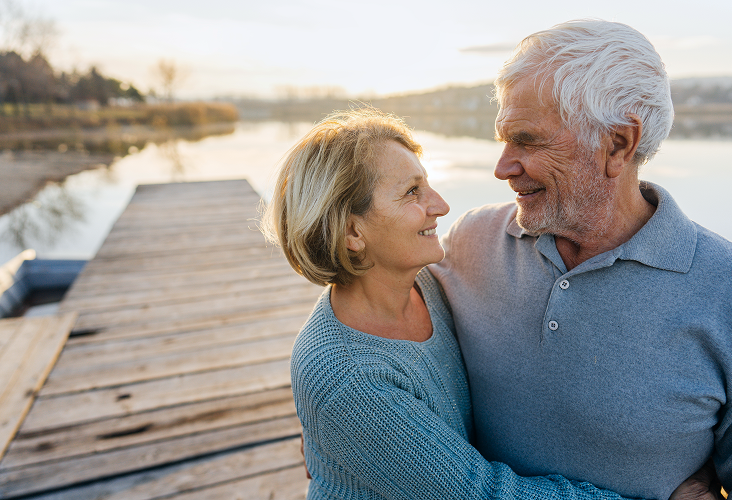 A happy elderly couple gazing into each other's eyes during sunset on a lakeside dock.