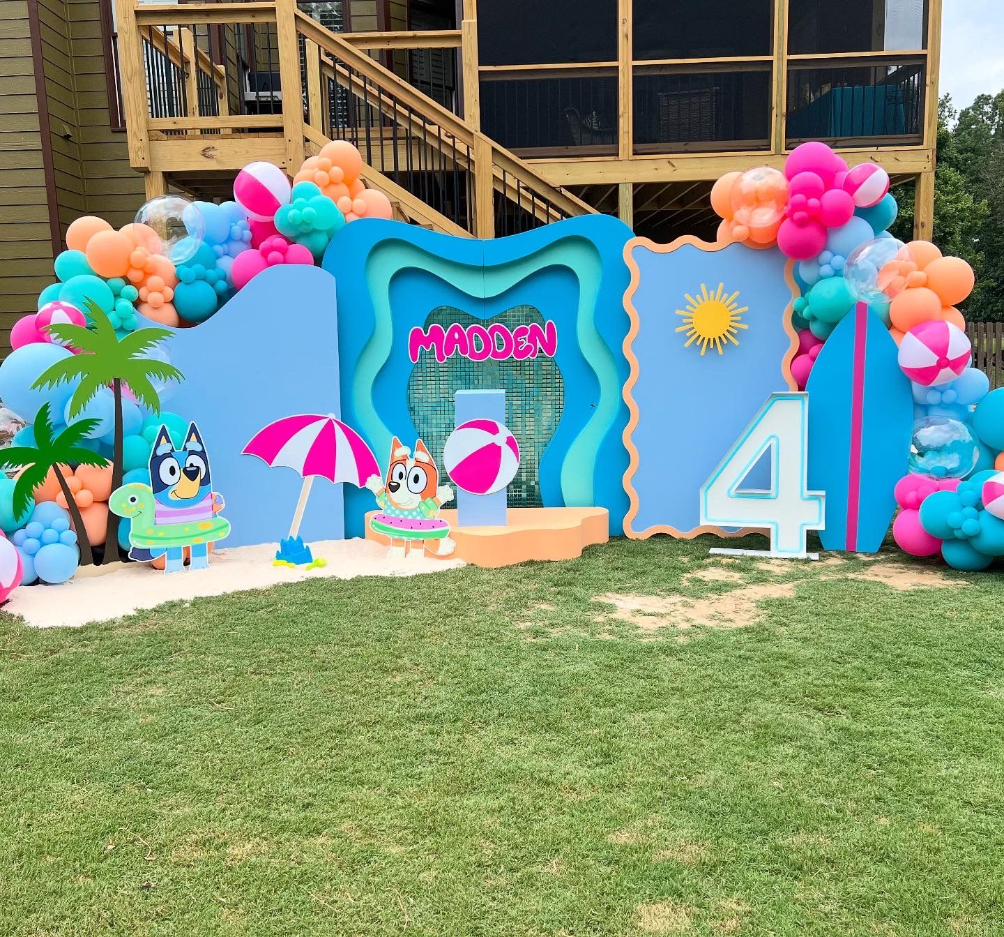Outdoor birthday backdrop with beach theme featuring colorful balloon garlands, palm tree, surfboard, sun accents, cartoon characters, and layered panels displaying Madden name and large number four celebration setup area.