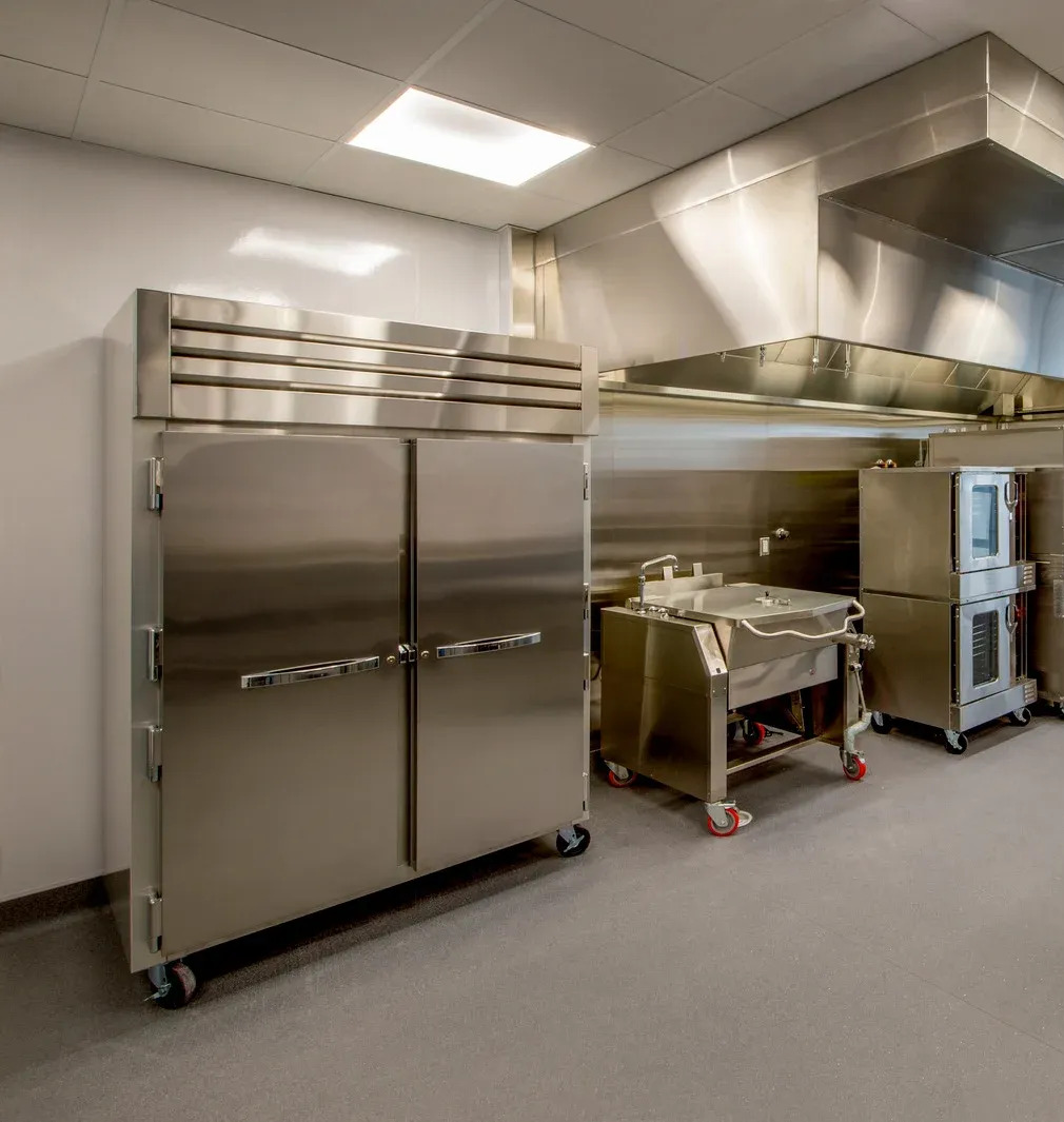 A pristine commercial kitchen featuring a large stainless steel double-door reach-in refrigerator. To the right, a tilting skillet and stacked convection ovens stand beneath a large metal exhaust hood.
