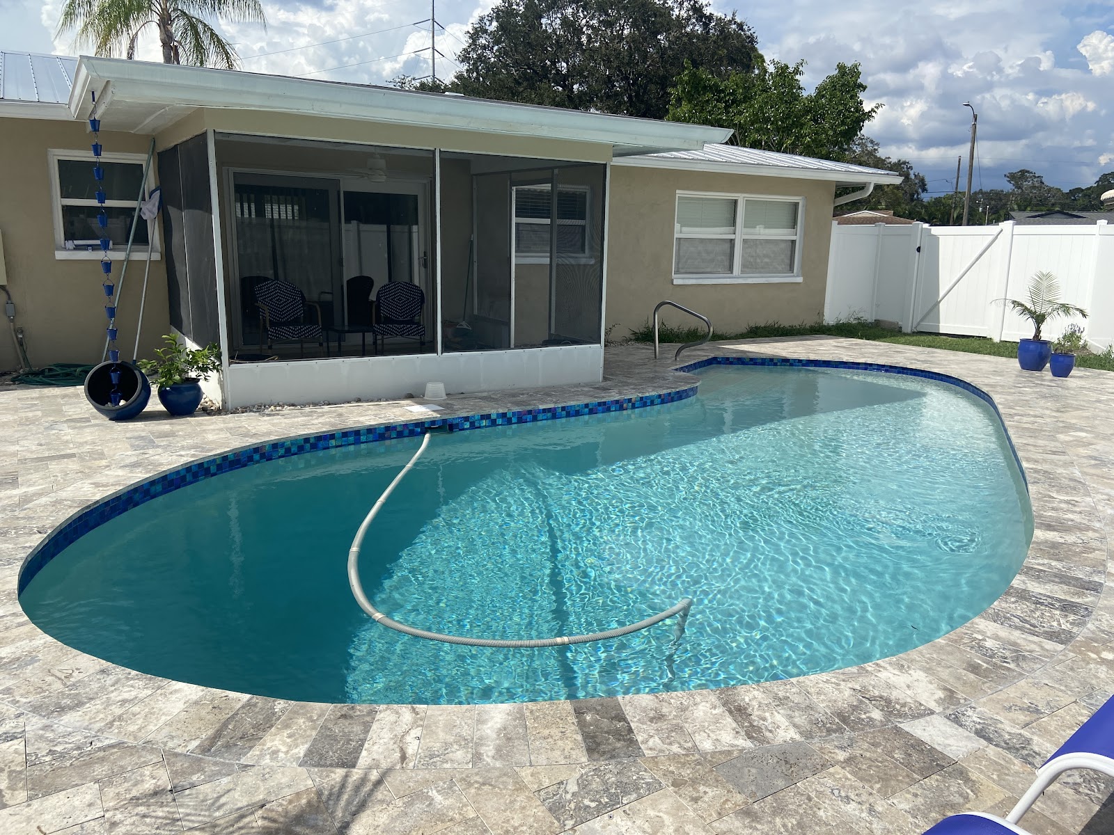 Freeform backyard swimming pool with clear blue water, light stone paver decking, and a pool vacuum hose in the water.
