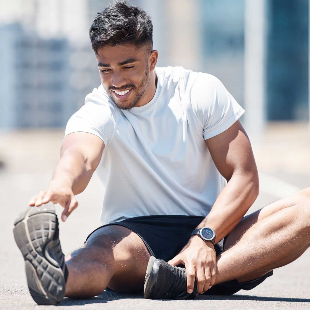 A man in workout clothes is stretching outdoors on pavement. He is wearing a light grey t-shirt, black shorts, and athletic shoes. He is reaching for his left foot with his right hand while his left leg is extended. He has a watch on his left wrist. In the background, there are out-of-focus buildings.