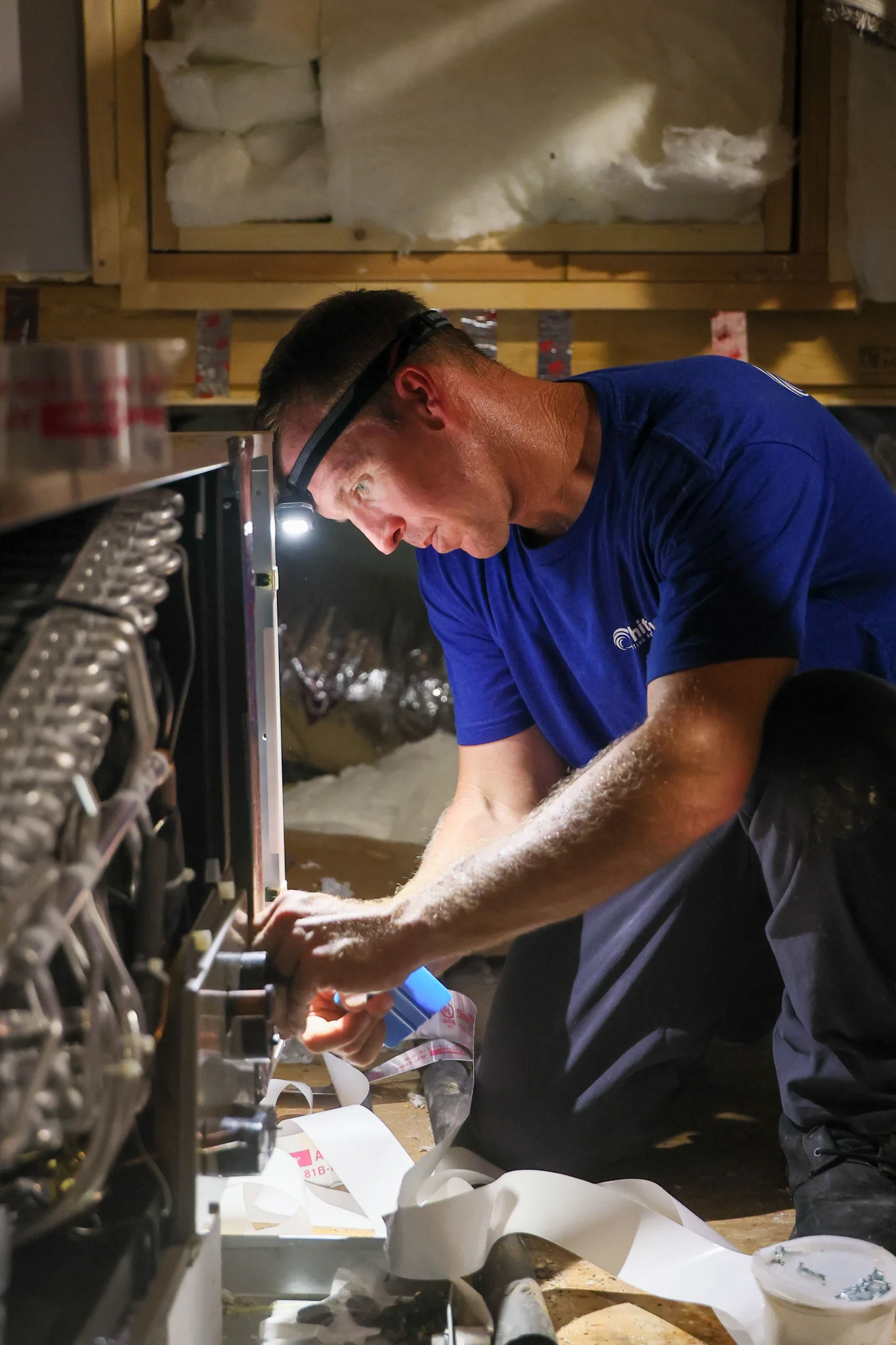 A man wearing a blue t-shirt and a headlamp is working on what appears to be an HVAC unit inside a c 