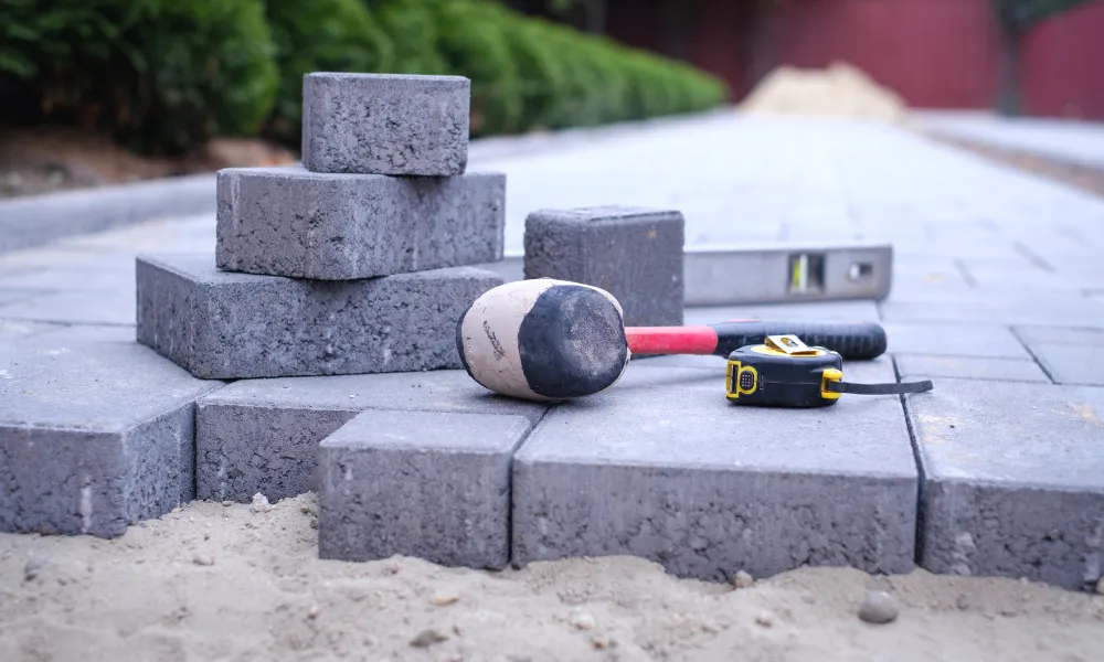 Construction tools including a rubber mallet, level, and tape measure are resting on a pile of gray concrete paving stones, with a blurred background of green bushes and a dirt pile. The scene suggests ongoing landscaping or construction work.