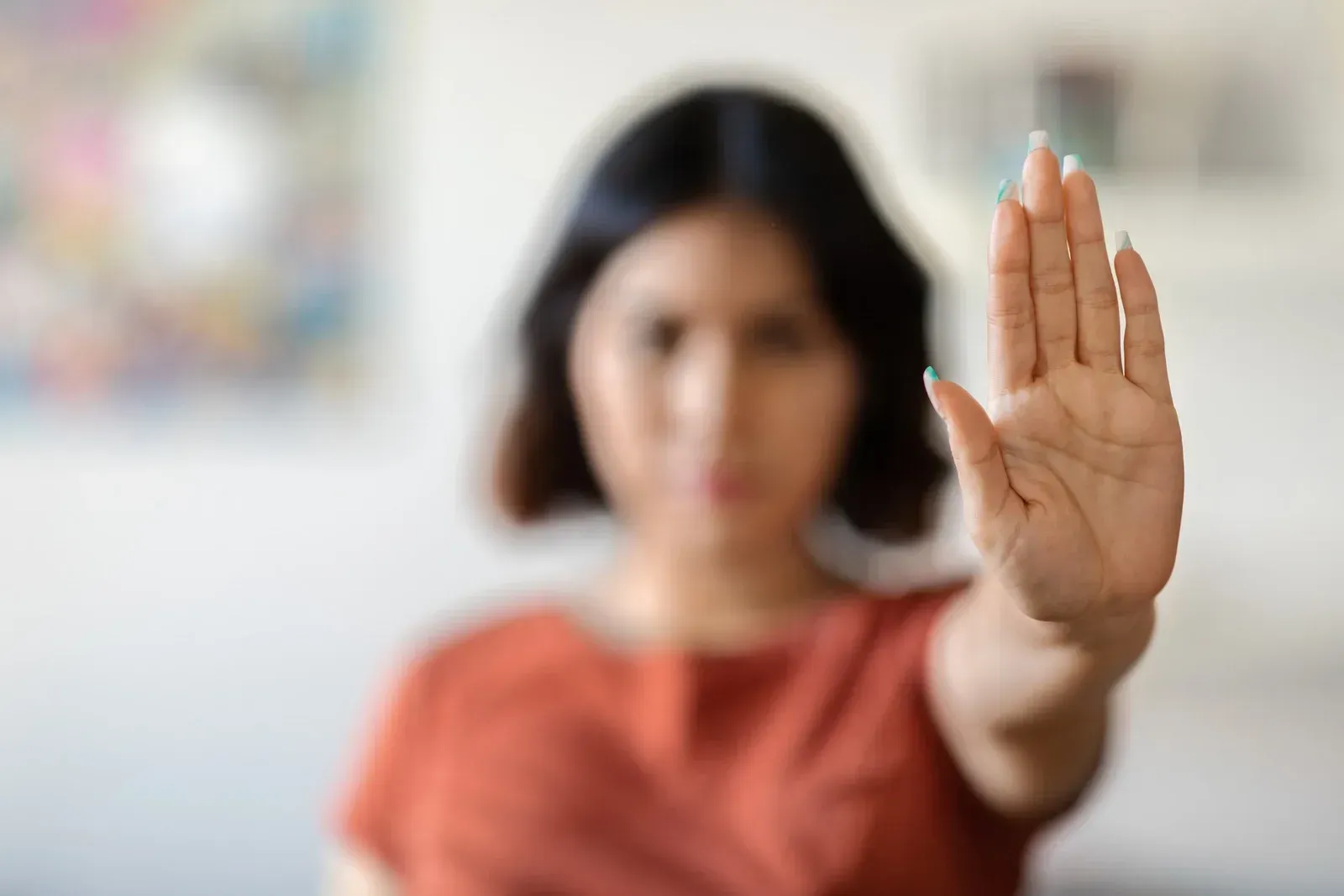 A person with short dark hair and wearing an orange-red shirt holds their hand up in a stop gesture, their palm facing forward. Their expression appears serious, and the background is softly blurred.