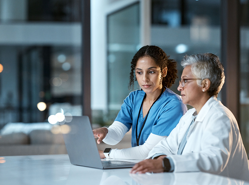 A woman in scrubs and a woman in a lab coat examining data on a screen.