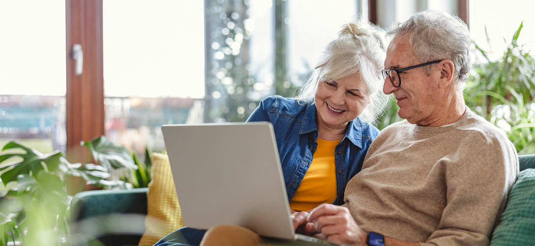 An elderly couple sitting on the couch looking at their laptop.