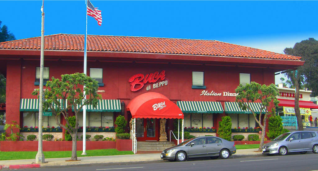 The exterior of Buca di Beppo Redondo Beach featuring the classic brick building shape, green and white striped windows, and an American flag.