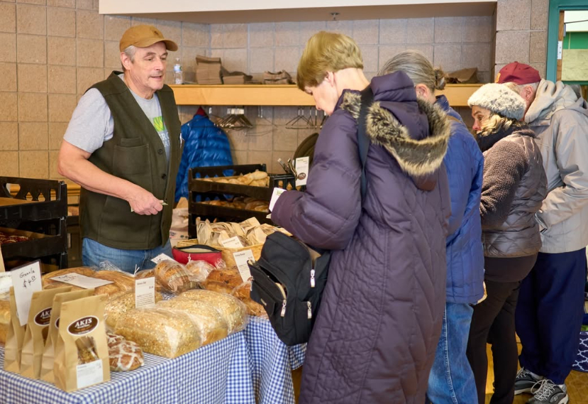 A local bakery vendor wearing a cap and vest chatting across a table with customers shopping for packaged artisan bread, loaves, and granola bags at an indoor market day.