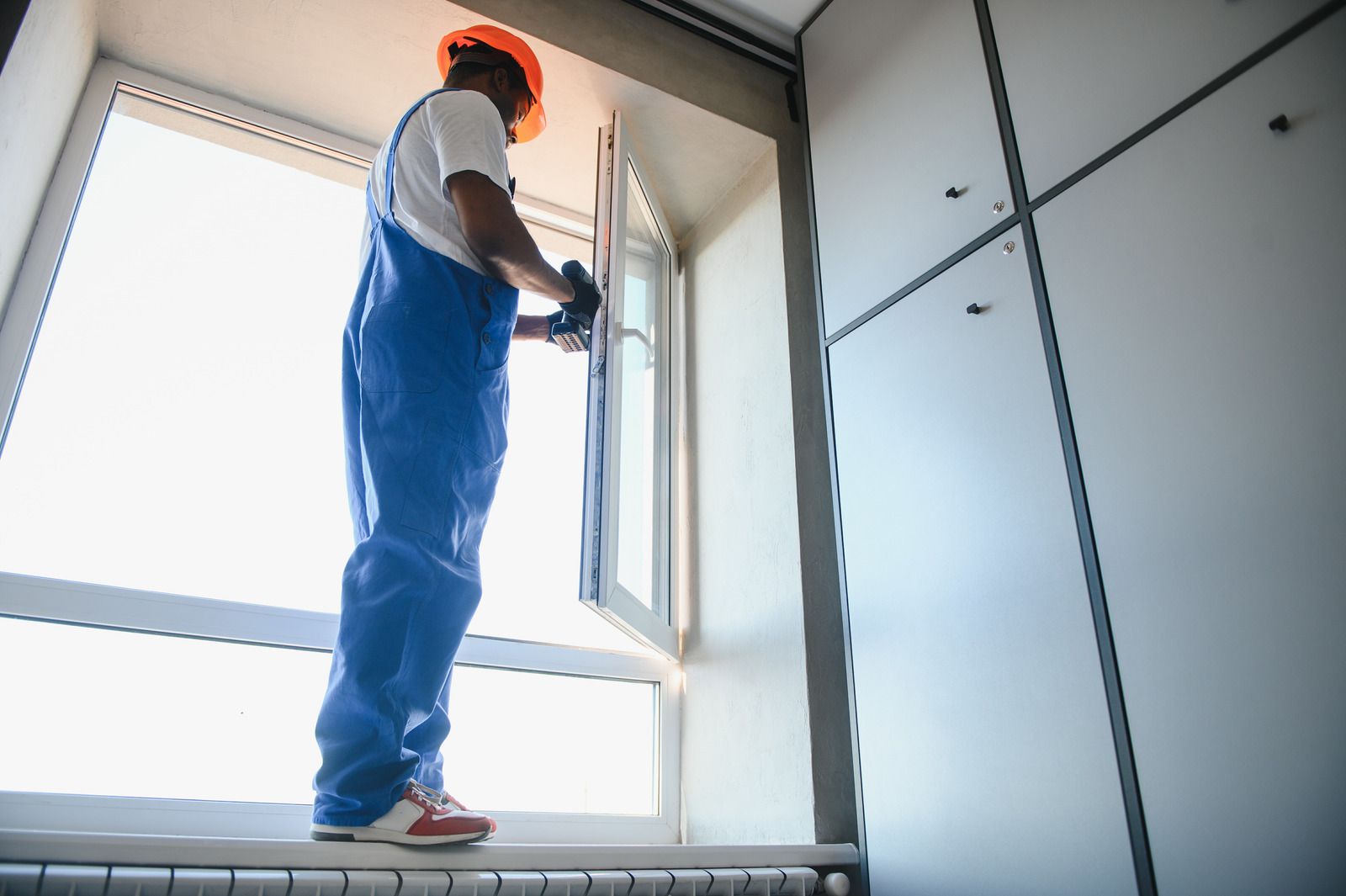 Window technician wearing blue overalls, gloves, and an orange safety helmet stands on a ledge using 