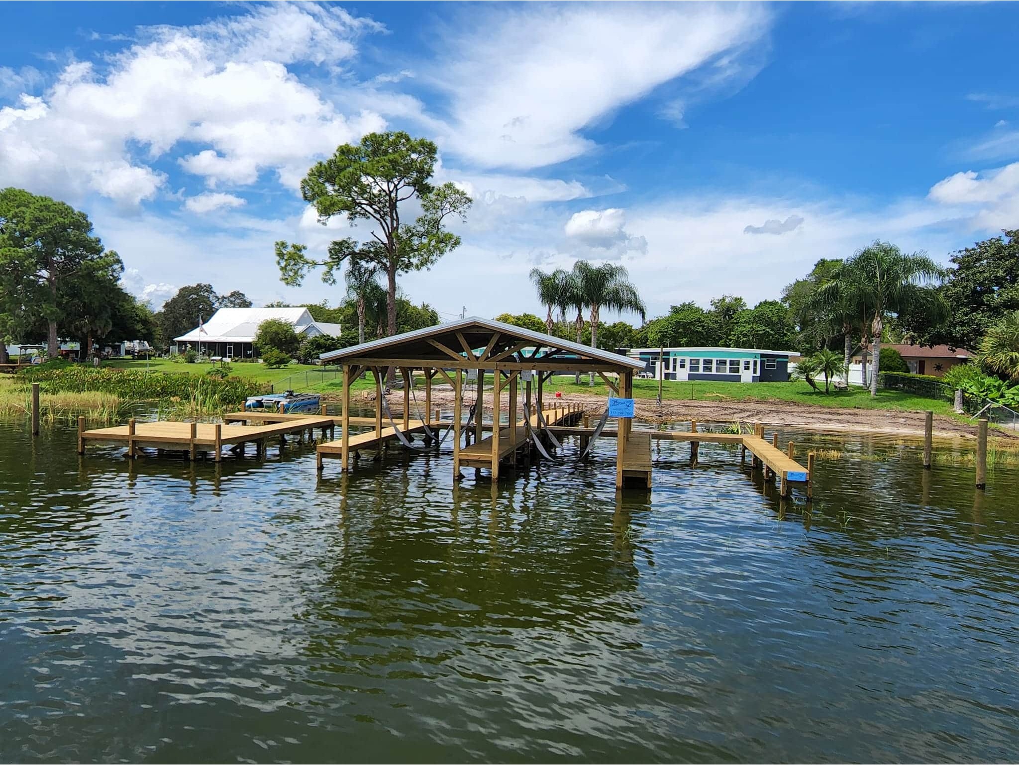 Calm lake scene with a wooden dock and boathouse. Lush greenery and palm trees line the shore, under a vibrant blue sky with fluffy clouds.