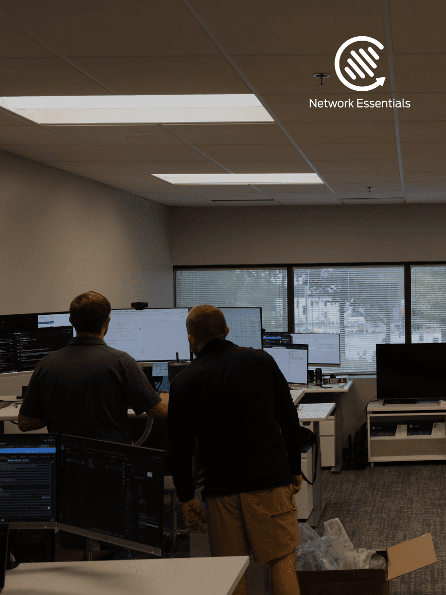 Two men in an office environment, with multiple computer monitors displaying code or data. The room features a drop ceiling with fluorescent lighting and large windows with blinds. A logo for "Network Essentials" is visible in the upper right corner.