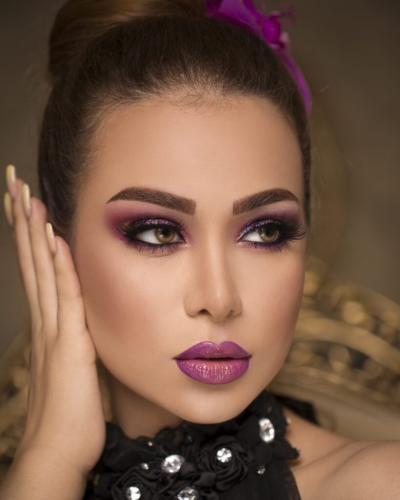 A close-up portrait of a woman with striking purple eye makeup and lipstick, her hair styled up and adorned with a purple flower. She is wearing a black, bejeweled collar and her left hand is gently touching her face.