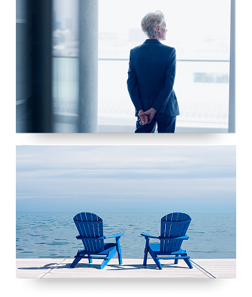 Short-haired woman wearing a suit staring out a large window with her hands crossed behind her back & two blue Muskoka chairs on a dock facing out towards the lake.