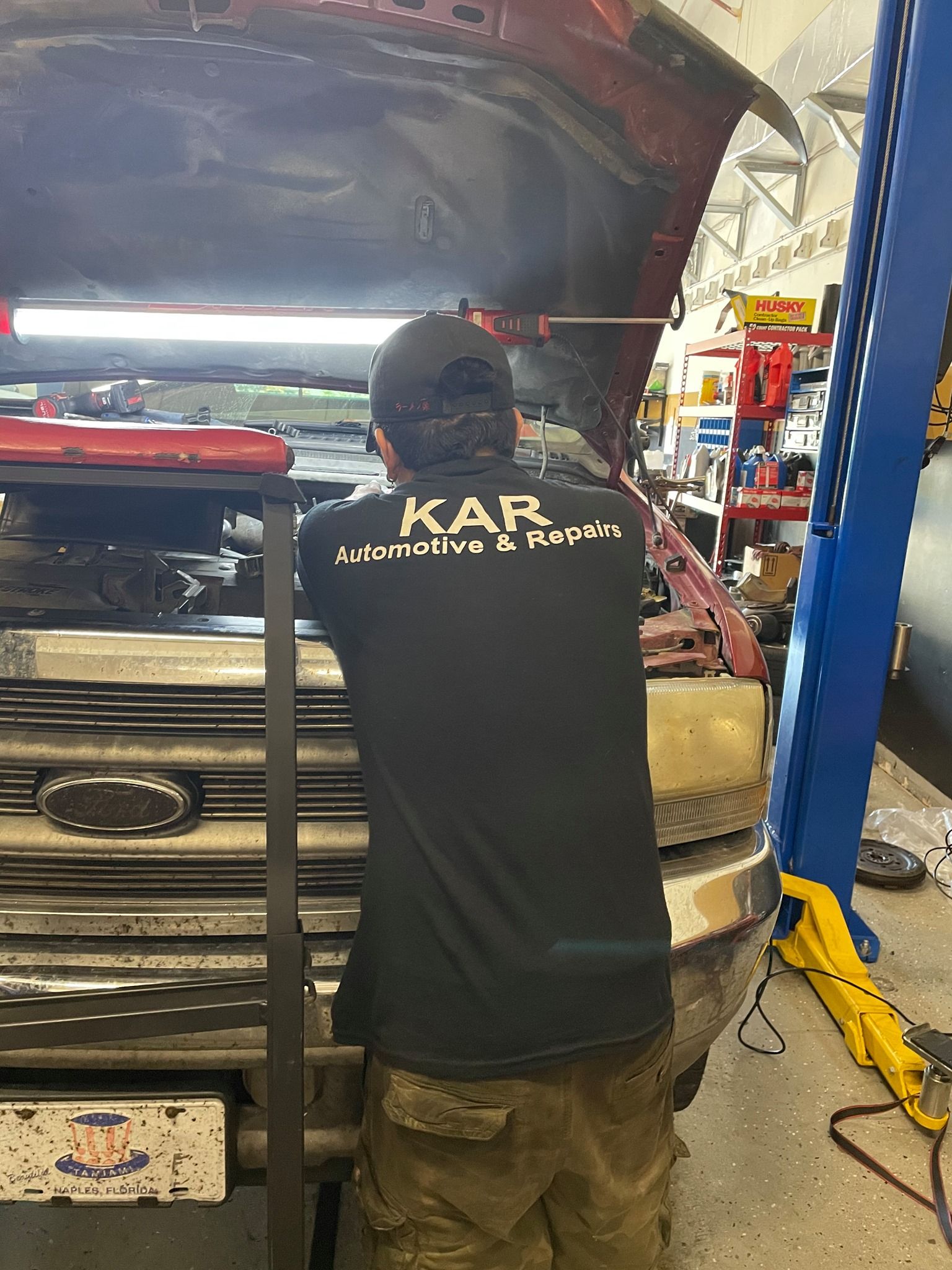 Mechanic wearing branded shirt working under the hood of a pickup truck inside a workshop. Tools, shelves, and equipment surround the area, showcasing hands-on vehicle diagnostics and repair in a professional automotive service environment.