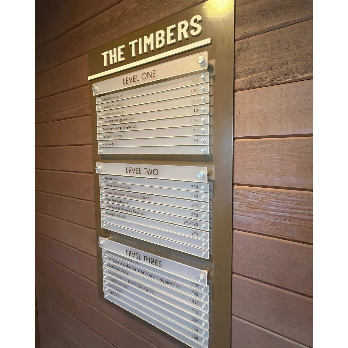 We recently completed this stunning custom Building Directory Sign for The Timbers, a multi-tenant office building located in beautiful Del Mar, CA.

A lobby directory needs to be two things: visually impressive and highly functional. For this project, we designed a premium directory that perfectly complements the building's warm, wood-plank interior architecture.

The foundation is a sleek gold aluminum back panel, topped with raised dimensional letters spelling out "THE TIMBERS." To ensure the property management team can easily update the board as tenants change, we utilized frosted acrylic strips mounted with decorative silver standoffs. This modular design allows individual tenant names to be swapped out quickly and cost-effectively without replacing the entire sign.

The result is a sophisticated, easy-to-read directory that helps visitors navigate all three levels of the building while elevating the overall lobby experience.