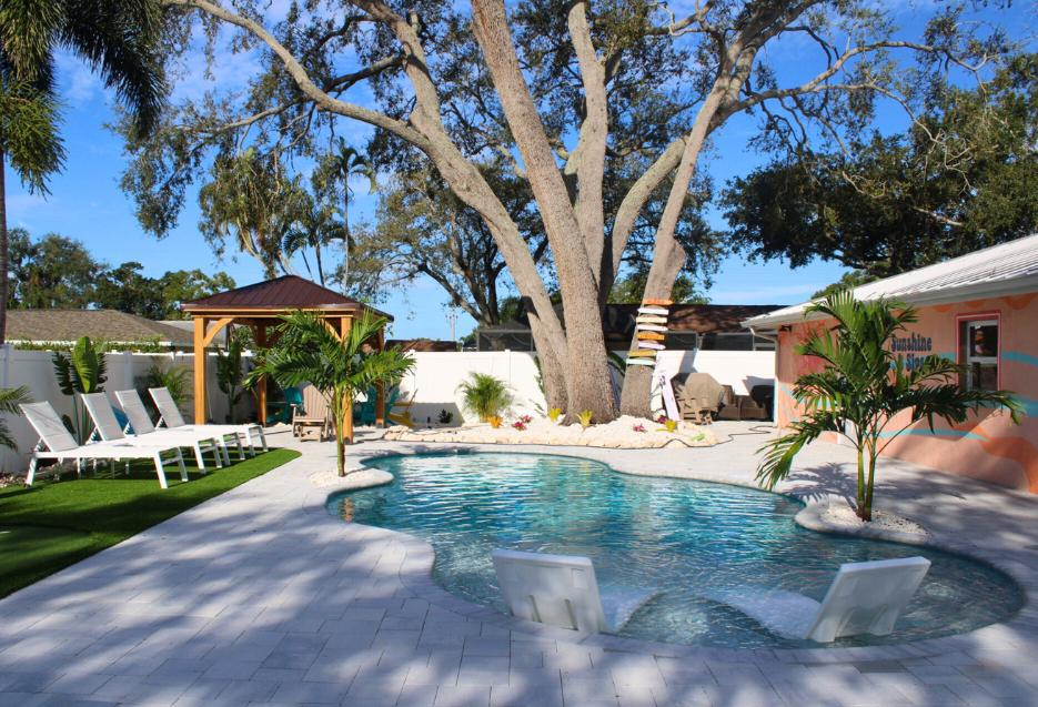 Residential backyard pool with clear blue water, light stone decking, lounge seating, and a large shade tree.