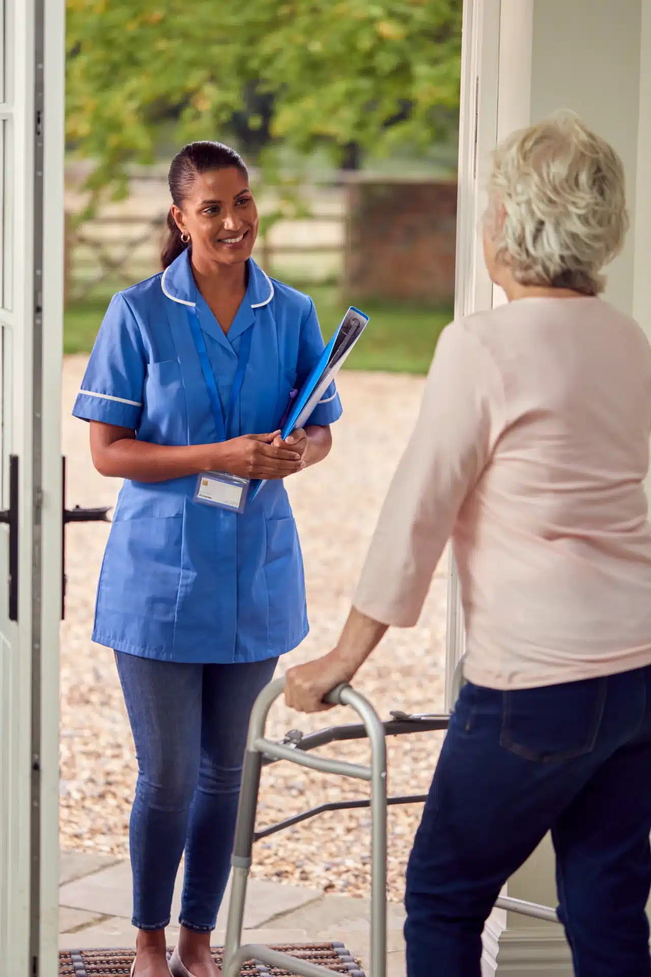 A female healthcare worker in a blue uniform and a name tag holds a folder and speaks to an elderly woman who is leaning on a walker. The scene appears to be at the entrance of a home, with an open door and greenery visible in the background.