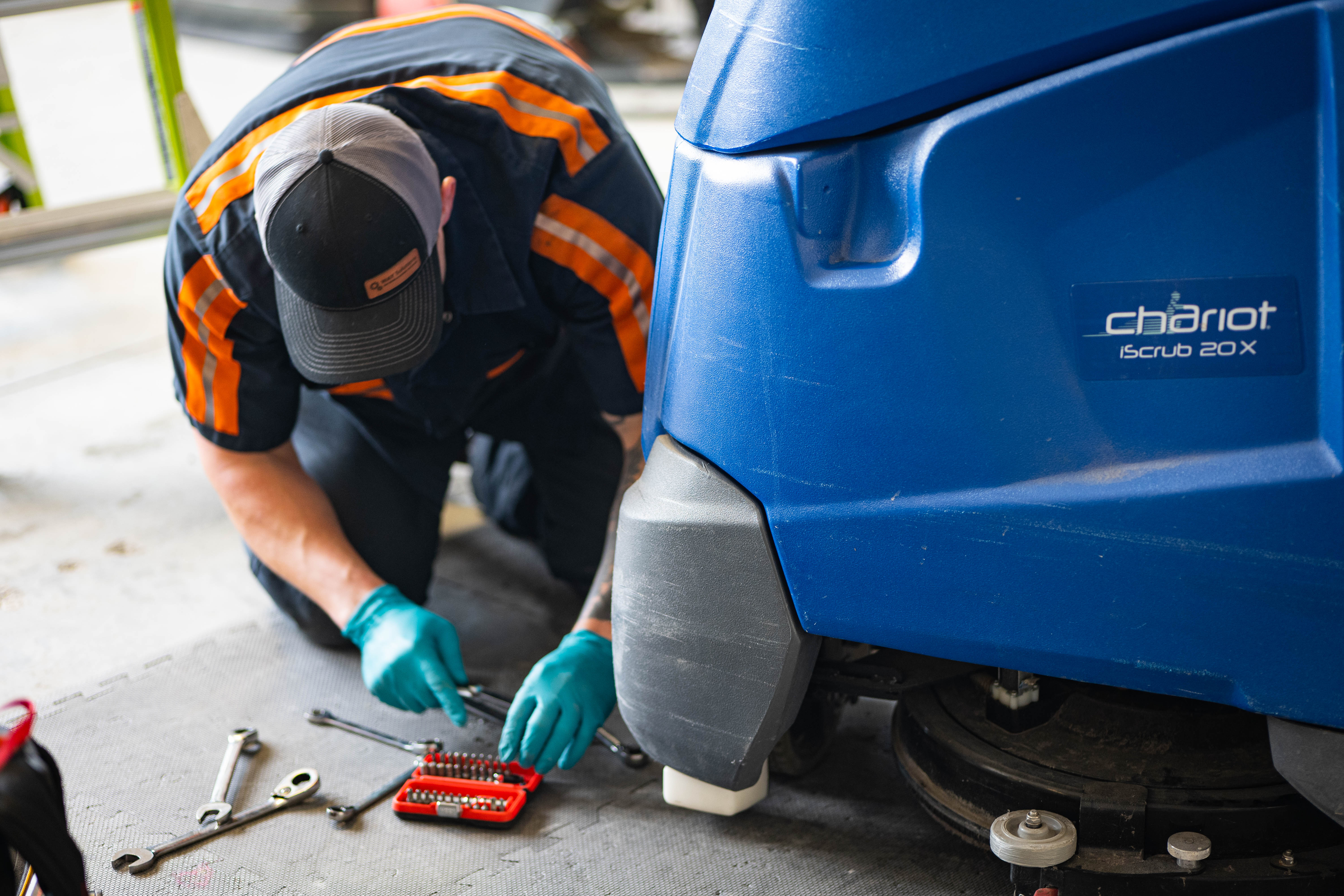 A professional WaySolutions technician performs detailed ground-level maintenance on the undercarriage of a blue Kärcher Chariot iScrub 20X stand-on floor scrubber in Layton, Utah. Our experts utilize professional hand tools to ensure all mechanical components and hardware are secure and ready for heavy-duty commercial cleaning.