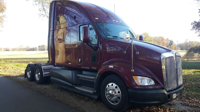 A large maroon Kenworth T700 sleeper semi-truck is parked on the edge of a paved road under a clear  