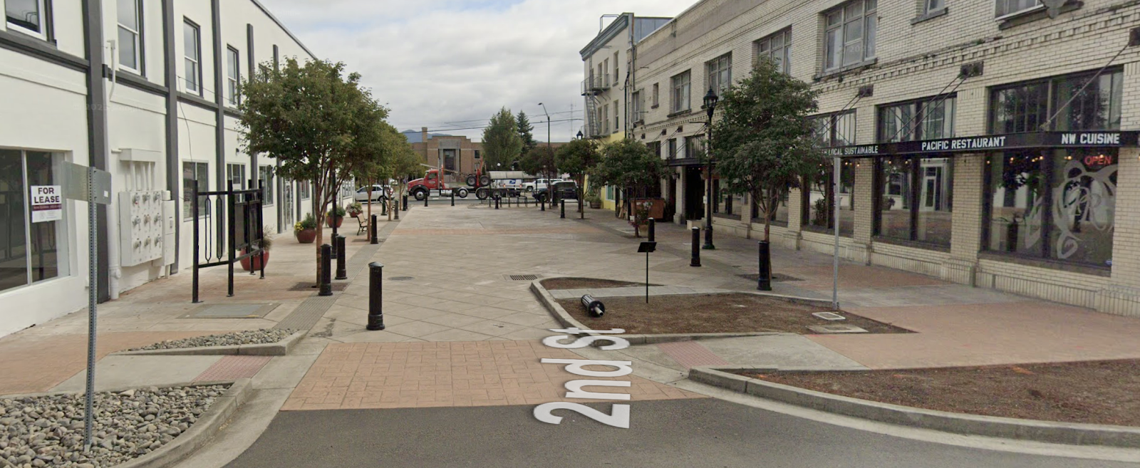 A wide view of a pedestrian street in a town with storefronts on either side. A "FOR LEASE" sign is visible on the left side of the street, and a restaurant with "PACIFIC RESTAURANT" and "NW CUISINE" signage is visible on the right. The street is paved with concrete and decorative tiles, and there are trees and benches along the sidewalks. A red truck is parked in the middle of the street.