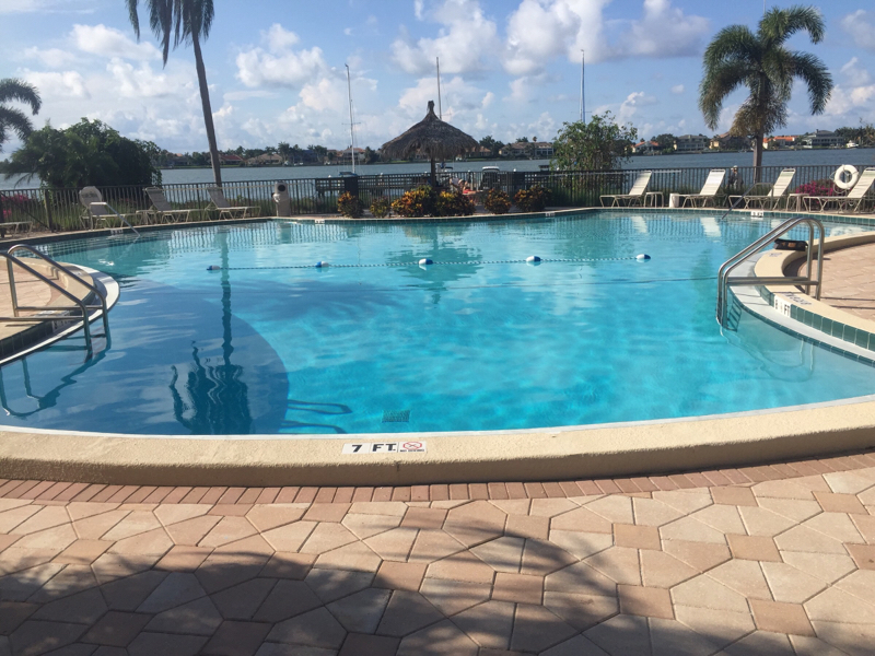 Large resort-style community swimming pool with blue water, palm trees, stone decking, and surrounding lounge areas.
